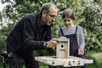 A man and girl look at a homemade bird box on a small table with trees in the background