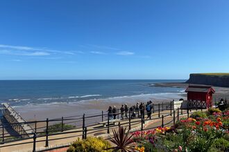 A group of people with binoculars looking out to sea. Saltburn's red Victorian cliff lift can be seen beside them, with the pier and Huntcliff in the distance