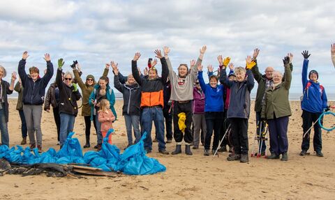 A group of men, women, one child and a baby in a row on a sandy beach in front of many blue bags of litter, most with arms in the air, smiling