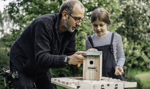 A man and girl look at a homemade bird box on a small table with trees in the background