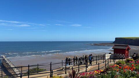 A group of people with binoculars looking out to sea. Saltburn's red Victorian cliff lift can be seen beside them, with the pier and Huntcliff in the distance