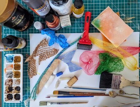 An artist's materials table showing a paint palette, coffee jar, paint bottles, paintbrushes, fruit nets and paper on a gridded table