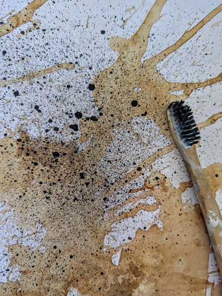 A toothbrush with wooden handle and black bristles lays upright on a white piece of paper marked with brown and black splashes and blown paint