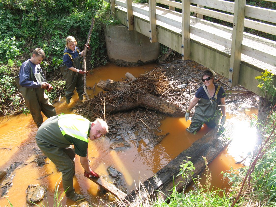 Volunteers clear debris washed down and trapped in a stretch of the river. They are working near a bridge, the river is orange from iron discharge and the volunteers are wearing waders. There are two women and two men working.