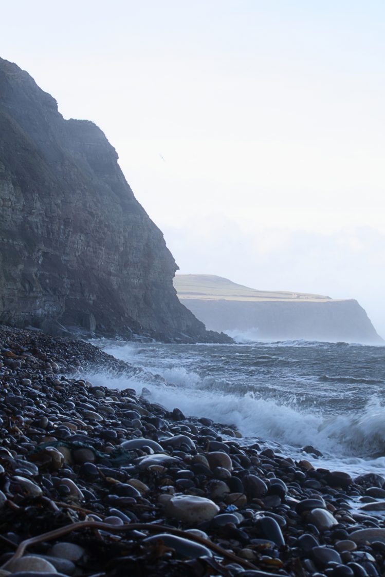 A view from the rock foreshore at Hummersea nature reserve. The tide is high and waves are crashing against the shiny, rounded stones. Cliffs rise form the sea and in the background, in sunlight, the fan-house just off the Cleveland Way is visible on the clifftop.